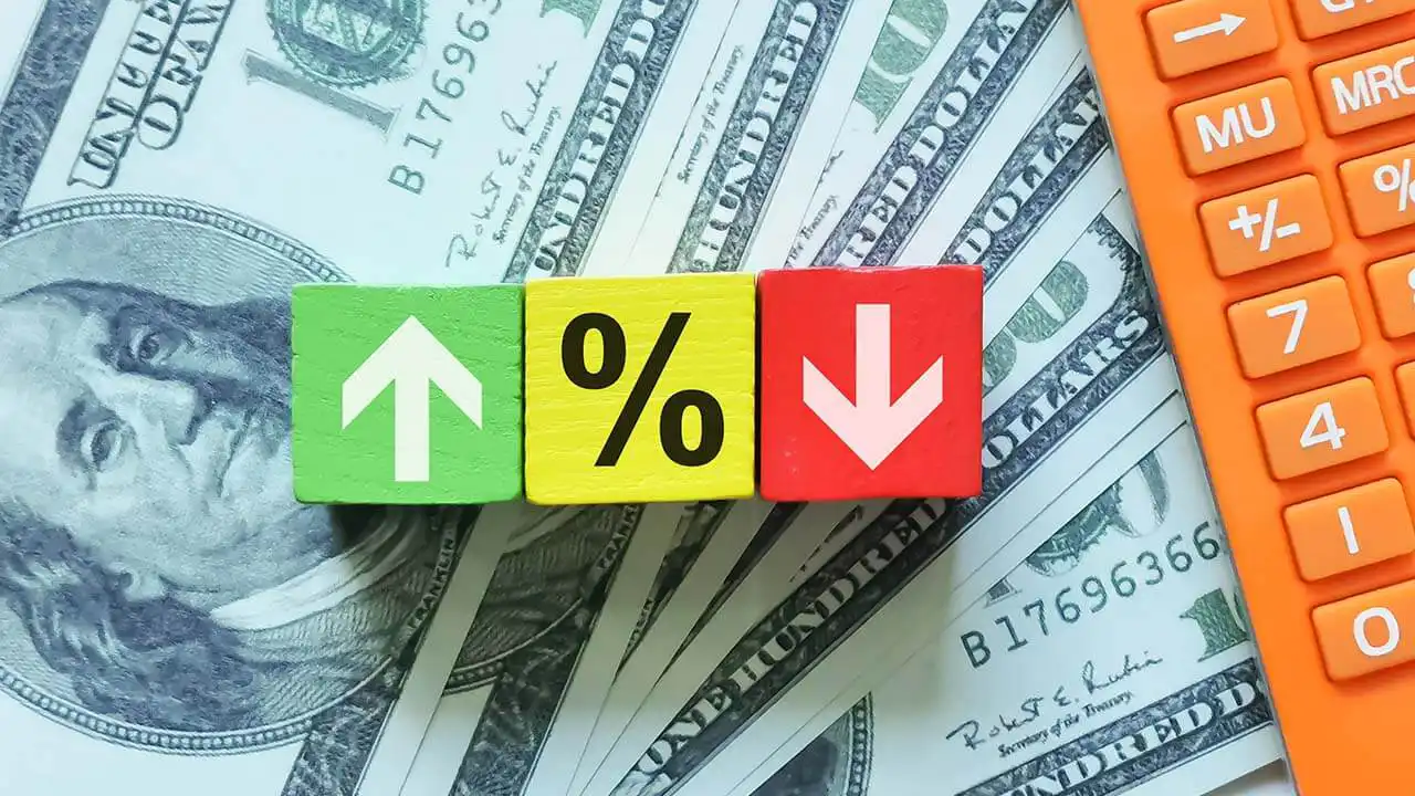 Image of apercentage icon and up and down arrow symbols on wooden cubes next to a calculator and atop $100 bills.